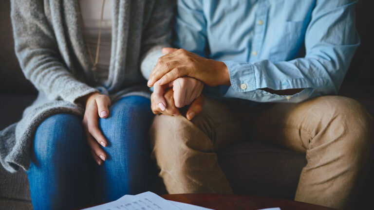 senior-couple-holding-hands-sitting-on-couch-with-life-insurance-policy-on-their-coffee-table
